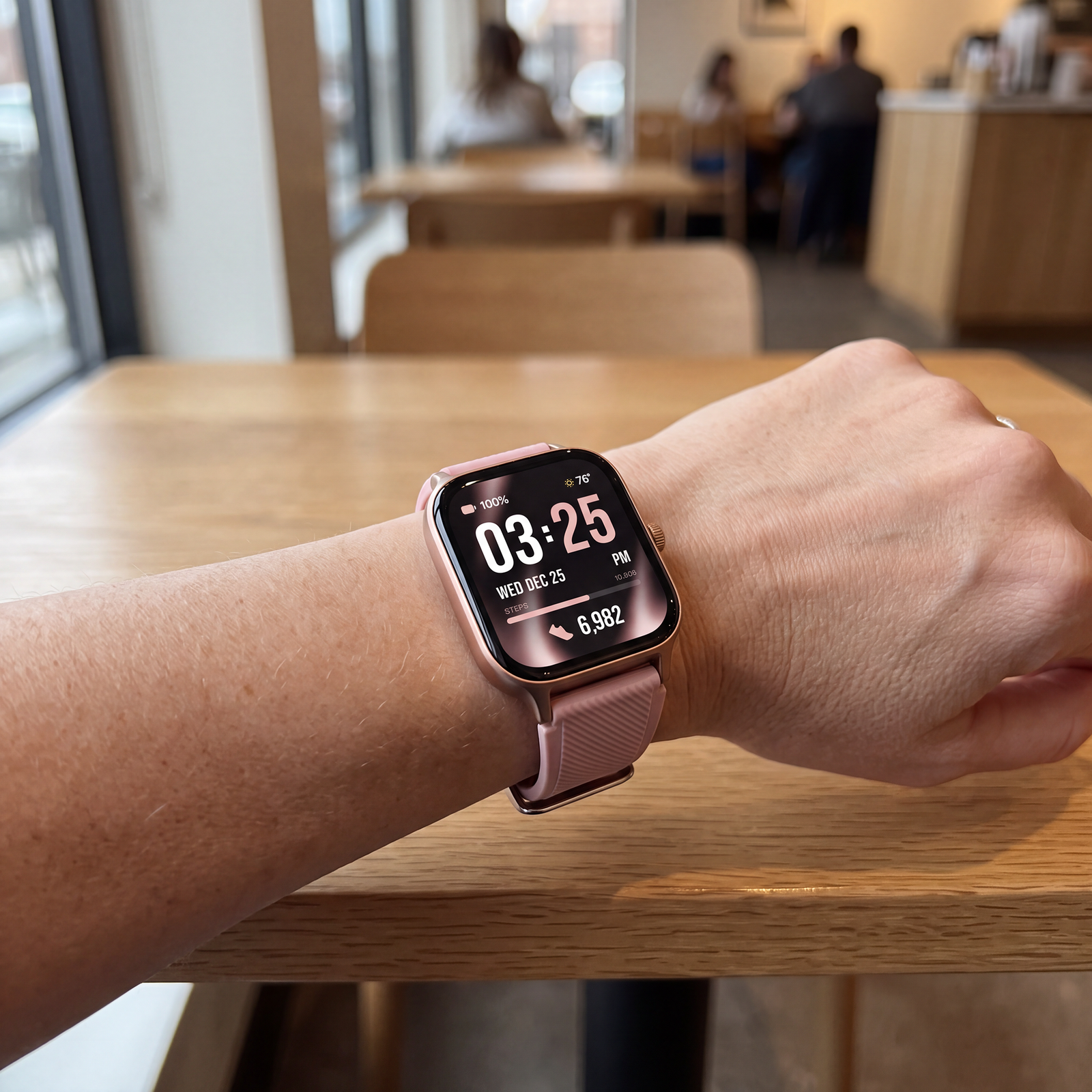 A smartwatch with a vintage rose band and rose gold-toned case worn on a person's wrist, resting on a light wood table in a blurred cafe setting.

Details